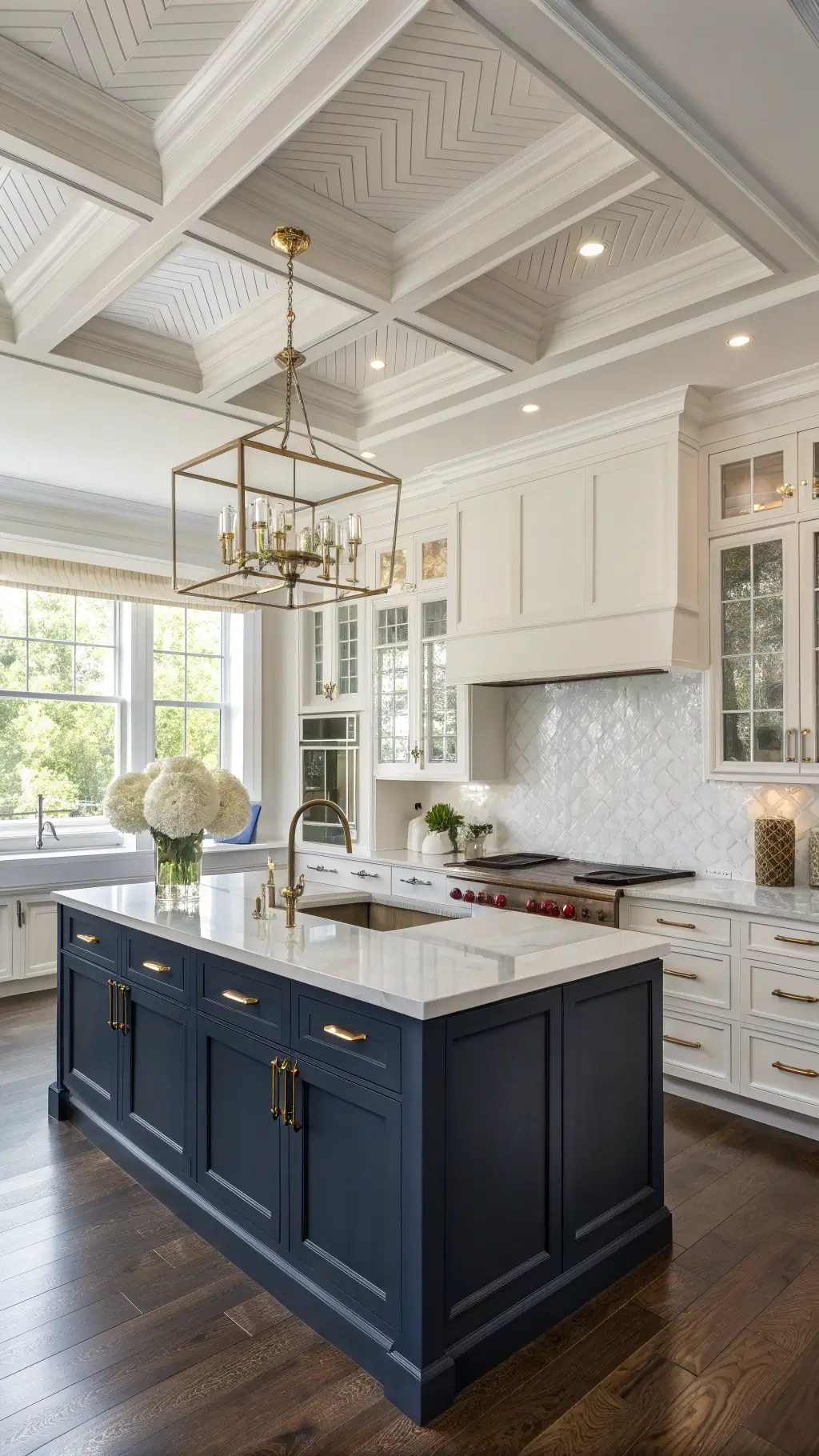 Elevated view of a transitional 15'x20' kitchen with chevron wood floors, coffered ceiling, navy blue island, white cabinets, and mixed metal hardware under diffused mid-morning light through roman shades, accented by a crystal chandelier and styled with white hydrangeas, glass canisters, and marble serving boards.
