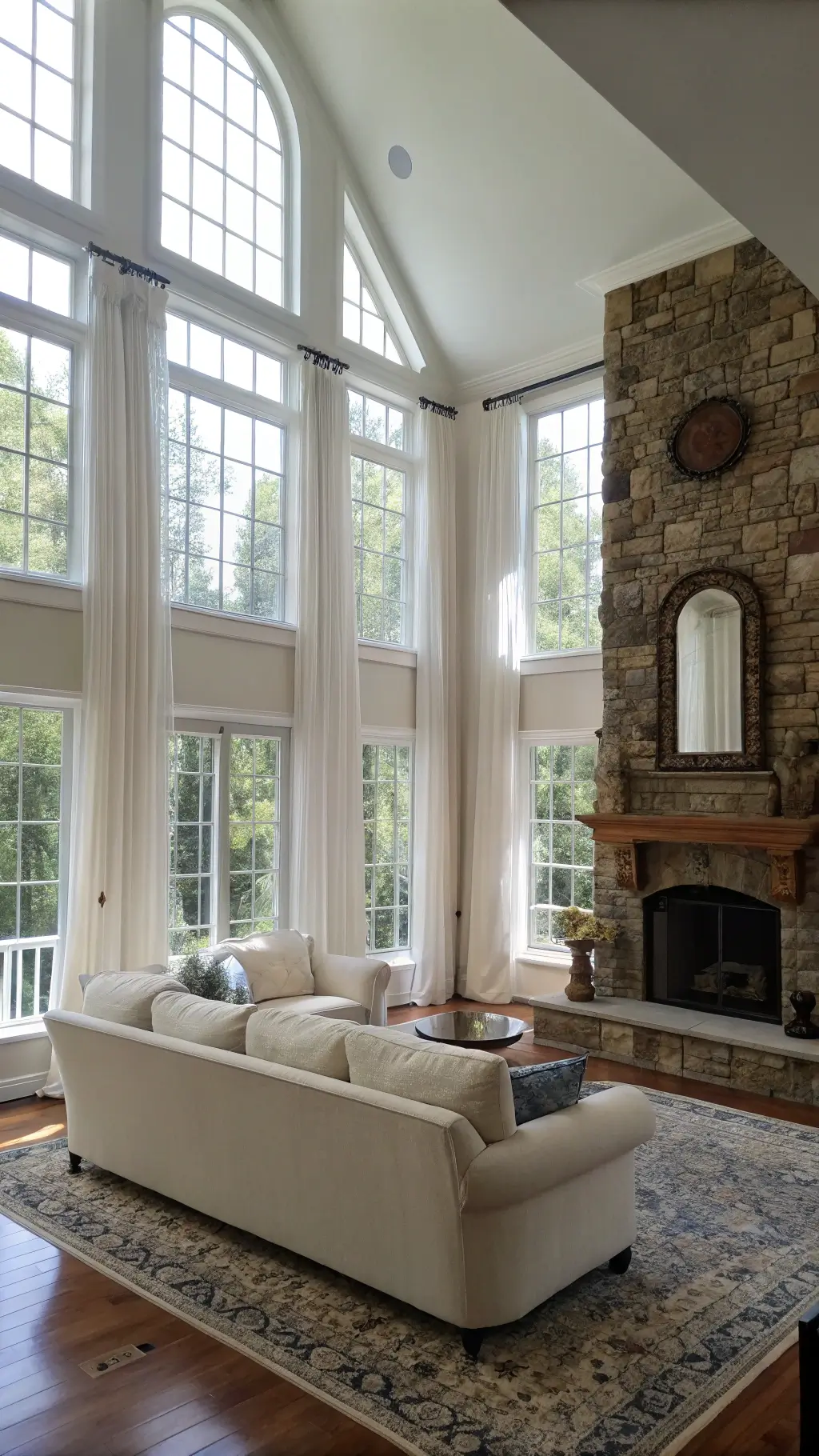 Spacious 20x24ft great room during mid-morning with double-height windows draped in gauzy white curtains, a cream bouclé sofa facing a stone fireplace with driftwood accents, and a vintage faded blue Turkish rug in the bright, naturally lit conversation area.