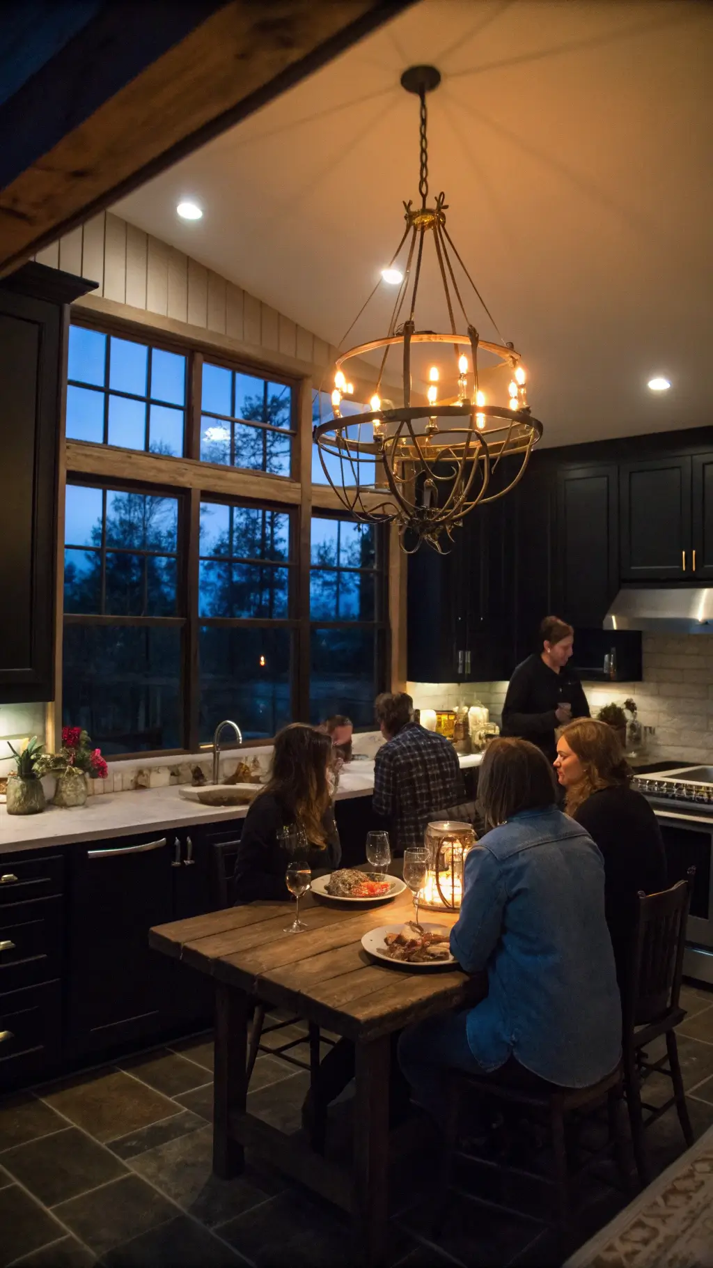 Bird's eye view of a spacious 18x20ft twilight kitchen with black weathered wood cabinets, an oversized iron chandelier, farmhouse table, and copper accents reflecting the evening light.